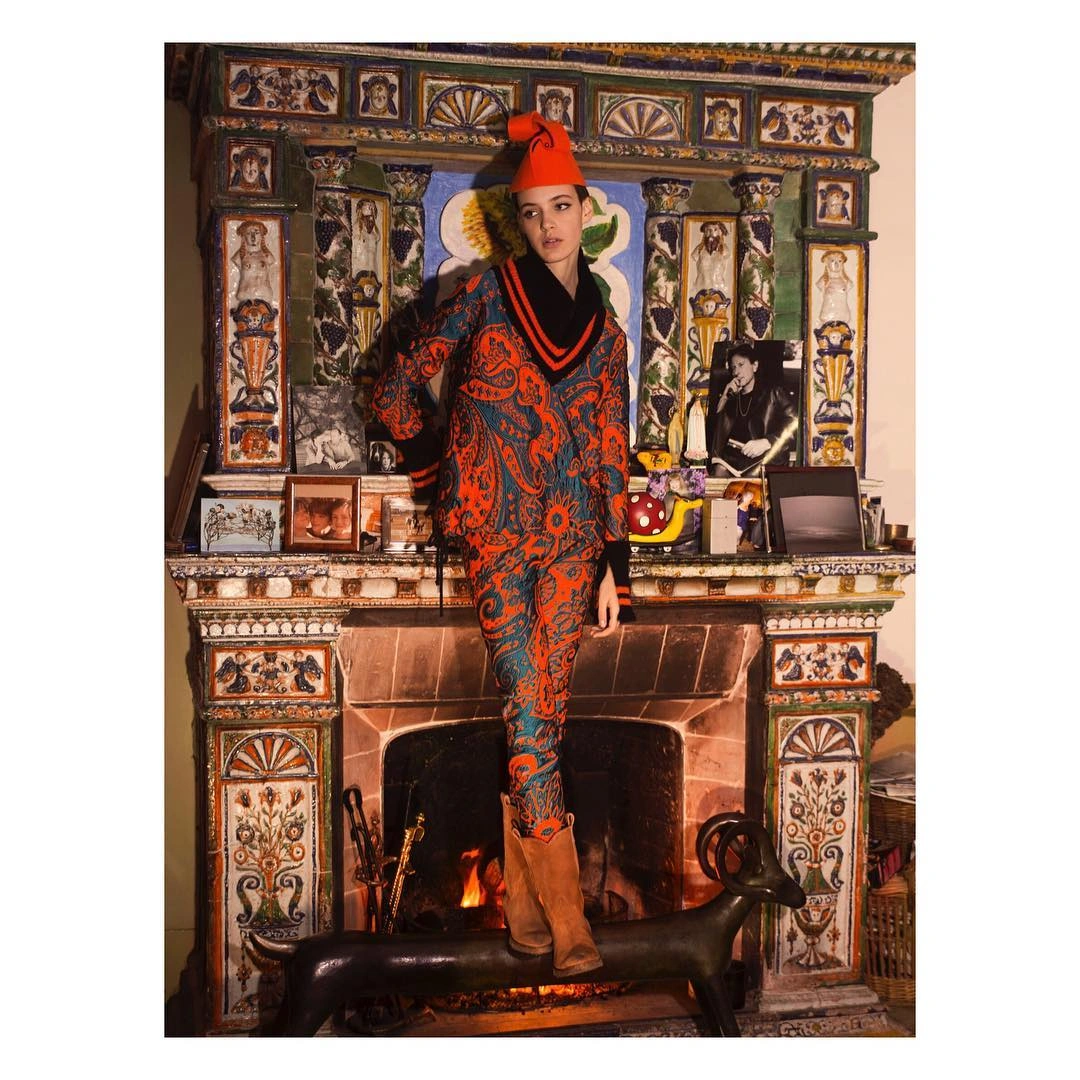 Model wearing orange patterned outfit and red hat standing in front of an ornate tiled fireplace by Simon Eeles