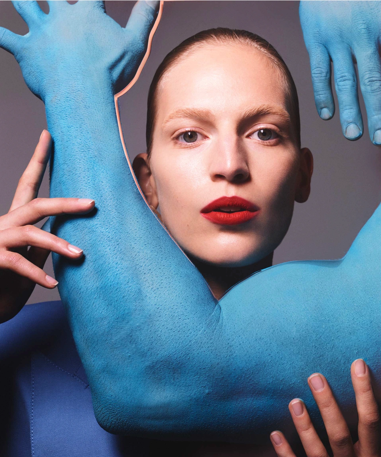 Close-up portrait of a woman with red lipstick framed by large blue-painted arms and hands against a grey studio backdrop.