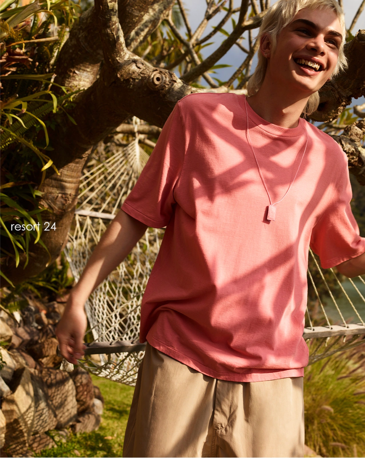 Young man smiling outdoors wearing a coral t-shirt and beige shorts near a tree and white hammock
