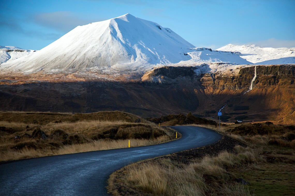 Scenic view of a winding asphalt road cutting through a grassy landscape toward a snow-covered mountain under a clear blue sky.
