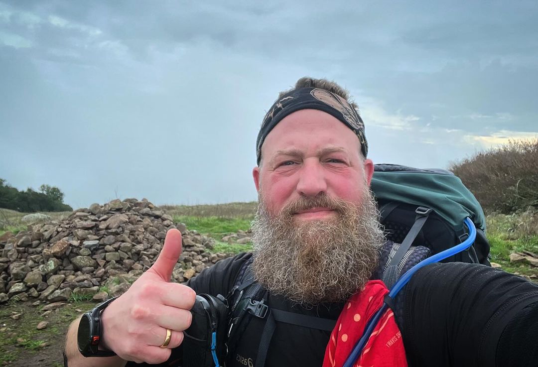 Man with beard wearing a backpack gives a thumbs up on a rural hillside with an overcast sky, wearing a black shirt and patterned headband.