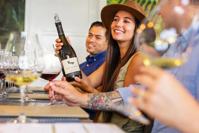 Woman in hat presents Sojourn wine bottle at a wine tasting with smiling guests, glasses, and vineyard views.