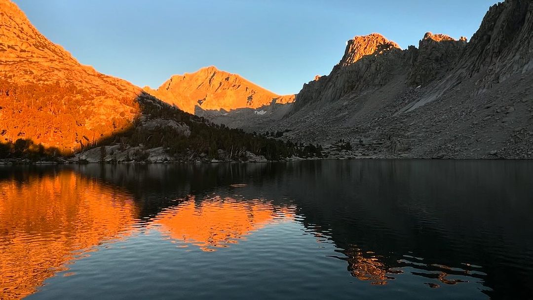 Scenic mountain lake at sunrise with the golden light reflecting on the water, surrounded by rocky peaks and sparse trees.