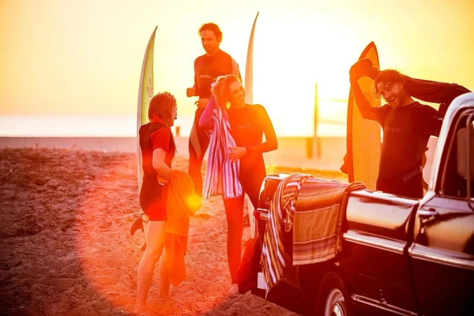 Group of surfers on sandy beach at sunrise near truck with surfboards, wearing wetsuits and towels. Golden light, ocean view.