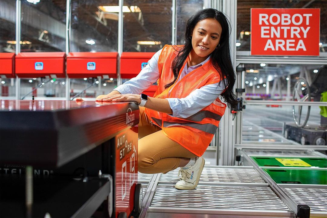 Young woman in safety vest crouching next to robot on rollers in industrial warehouse, smiling and checking settings near a Robot Entry Area sign.