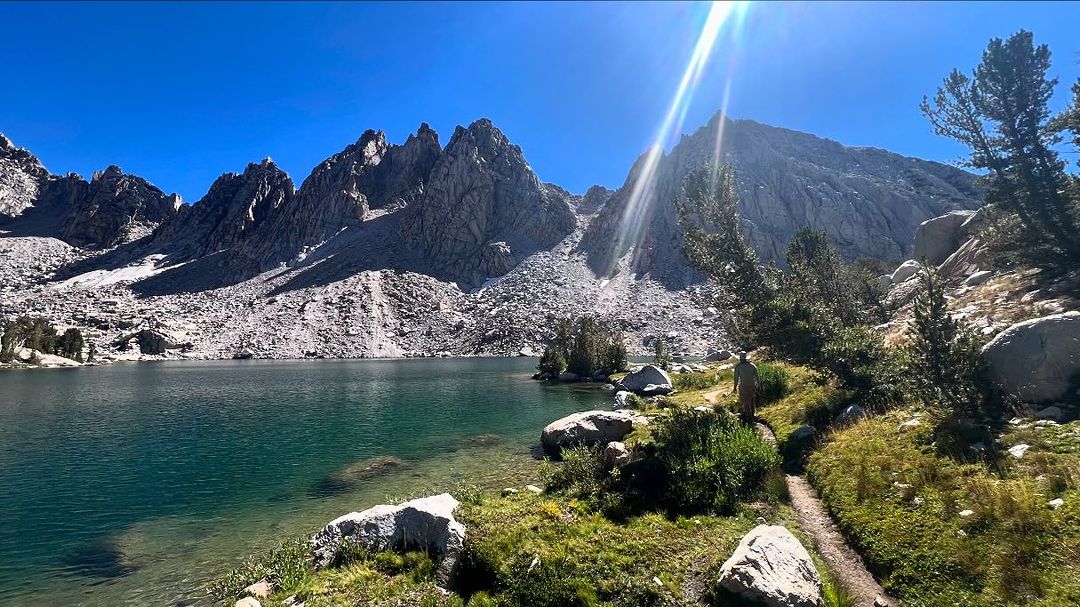 Scenic view of a mountain lake surrounded by rocky peaks and lush vegetation, with a person walking along a path on a sunny day.