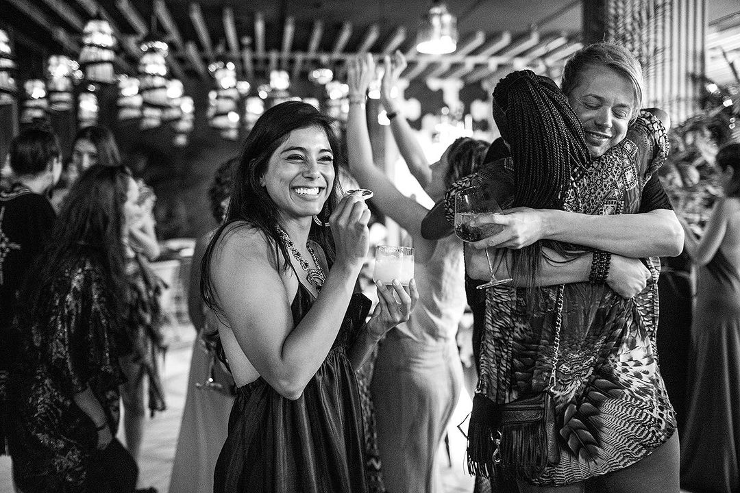 Black and white shot captures a smiling woman with a drink amidst a lively gathering, with a heartfelt embrace adding to the joyous scene.