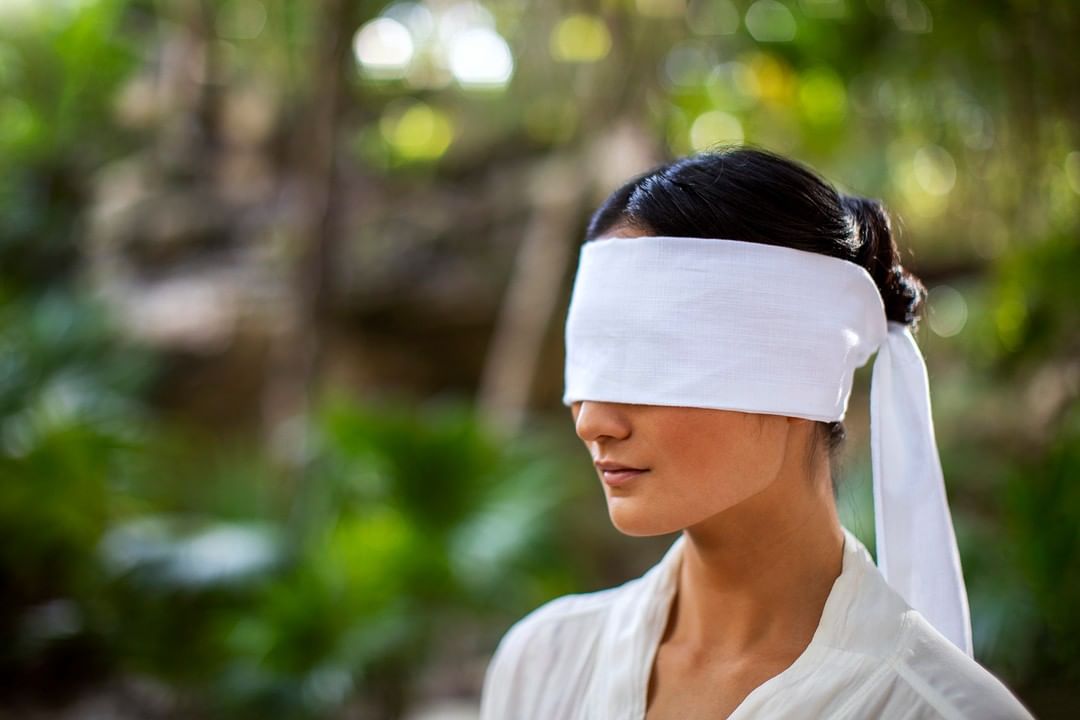 Woman with a white blindfold stands outdoors with a blurred green foliage background, creating a serene and enigmatic visual.