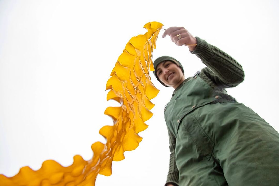 Woman in green overalls smiles while holding a large kelp leaf against a bright white sky in a low-angle shot.