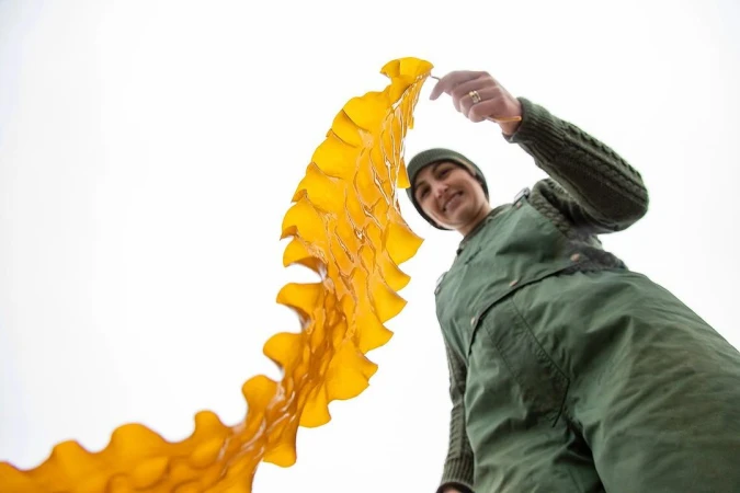 Woman in green overalls smiles while holding a large kelp leaf against a bright white sky in a low-angle shot.