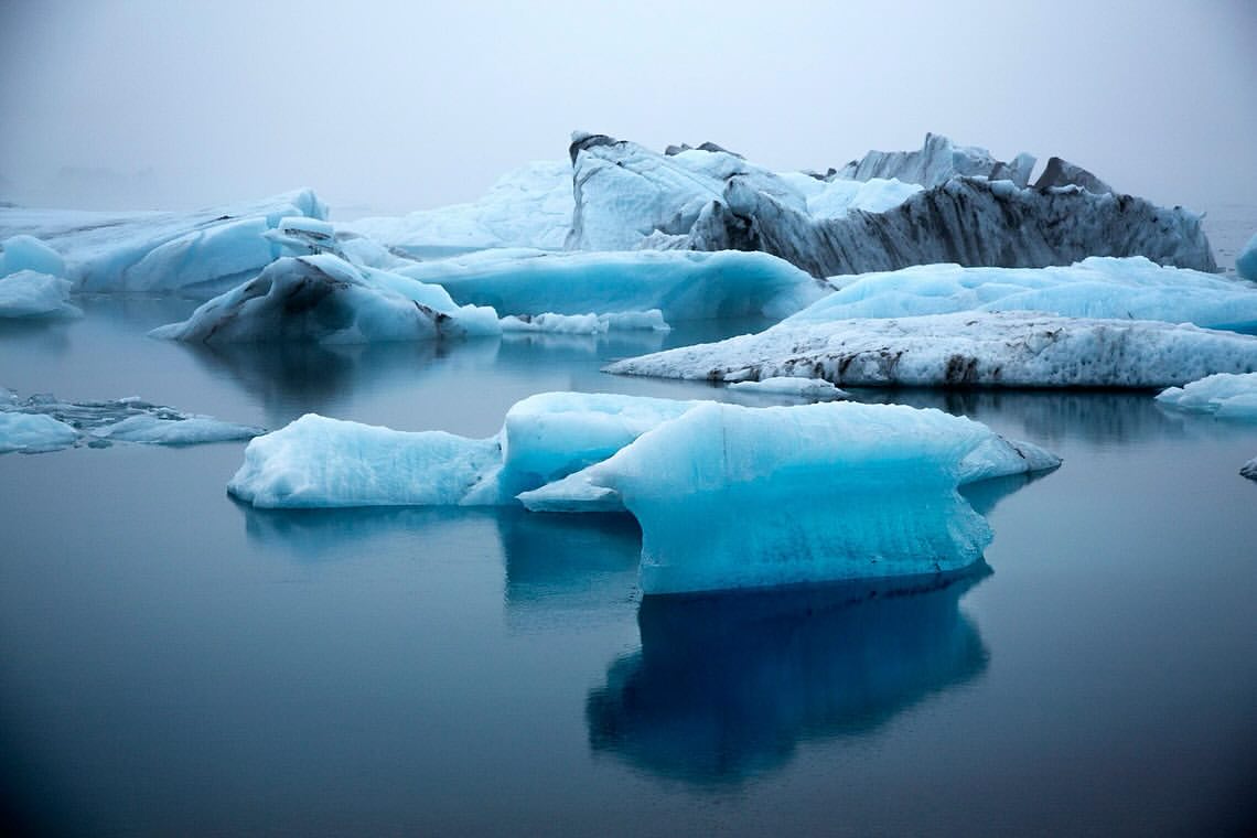 Icy turquoise-blue icebergs float in still water on a foggy day. Landscape view of natural ice formations with cool color palette.