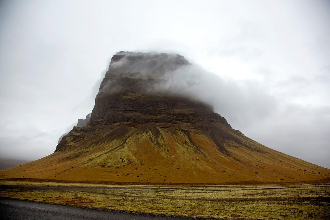 Scenic view of a brown mountain partially covered in low-lying clouds, under an overcast sky, in a natural landscape setting.