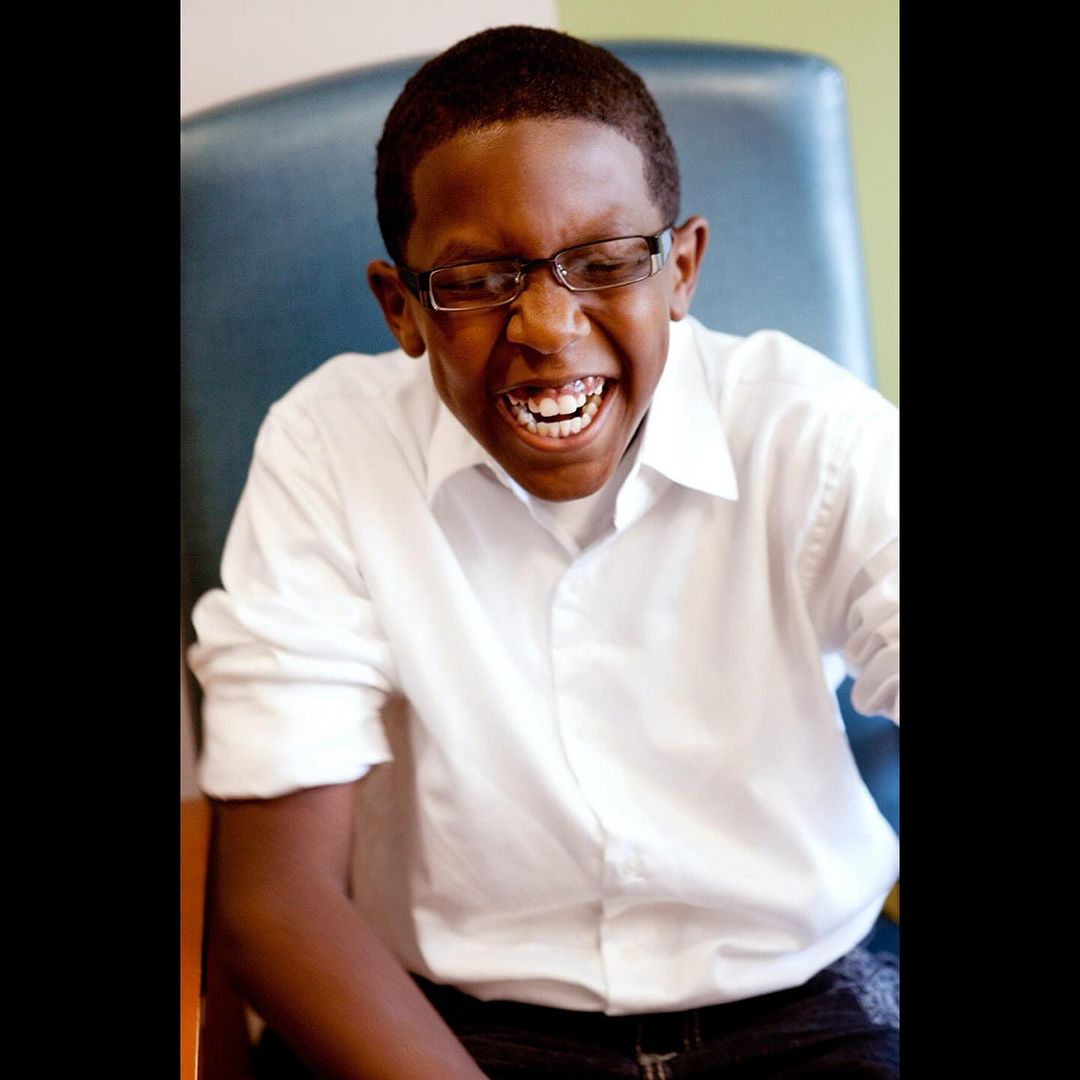 Portrait of a smiling young man with dark hair and glasses wearing a white button-down shirt sitting indoors in front of a green wall.