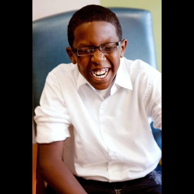 Portrait of a smiling young man with dark hair and glasses wearing a white button-down shirt sitting indoors in front of a green wall.