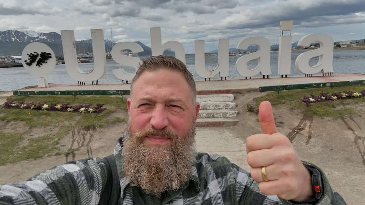 Bearded man gives thumbs up in front of 'Ushuaia' sign, set against a backdrop of a harbor, water, and snow-capped mountains.