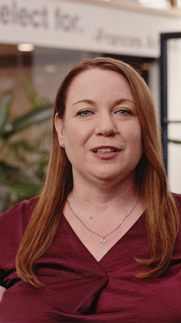 Portrait of an adult woman with auburn hair wearing a maroon top, standing in front of an office with greenery.