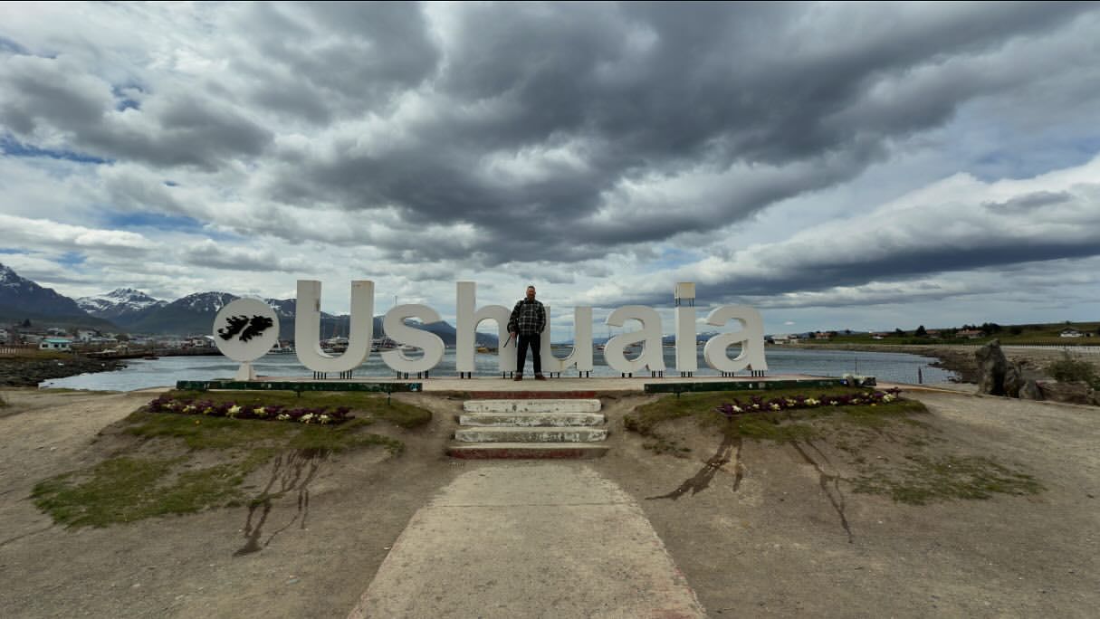 Adult stands before 'Ushuaia' sign with cloudy sky, mountains, and water backdrop. The setting showcases travel in Ushuaia.