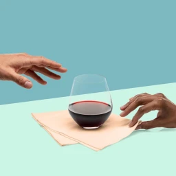 Two hands reaching towards a stemmed wine glass with red wine resting on a peach napkin against a two-tone background
