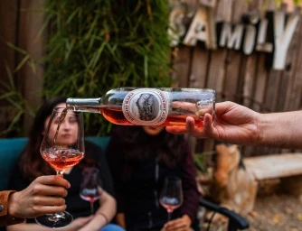 Hand pouring rose wine from a bottle into a stemmed glass, with people seated in an outdoor background.