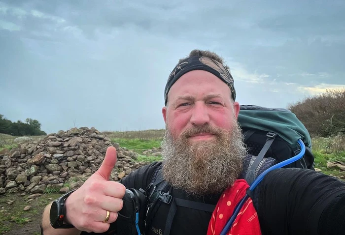 Bearded hiker wearing a dark shirt and backpack giving a thumbs up on an outdoor trail under an overcast sky.