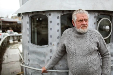 Elderly man with white beard wearing a grey cable-knit sweater standing on a ship deck by a metal structure