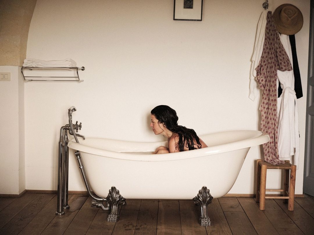 Woman in a clawfoot bathtub in a white-walled bathroom, with a wooden floor, vanity, and clothing rack, bathed in natural light.