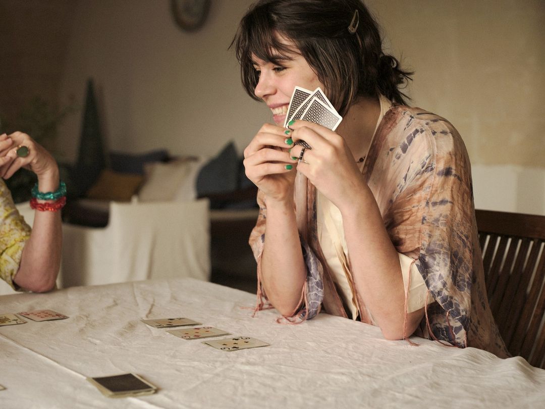 Two women are playing cards indoors, with one smiling while holding cards and the other's arm visible on the table with playing cards.