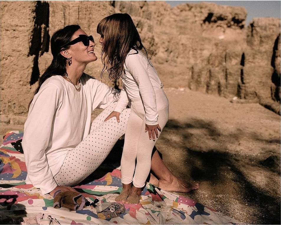 Woman and child enjoy a sunny outdoor moment on a patterned blanket near a rock formation, both in white outfits, sharing a joyful expression.