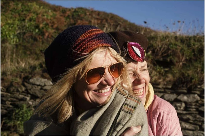 Two smiling women outdoors wearing winter hats, sunglasses, and layered scarves against a stone wall background.