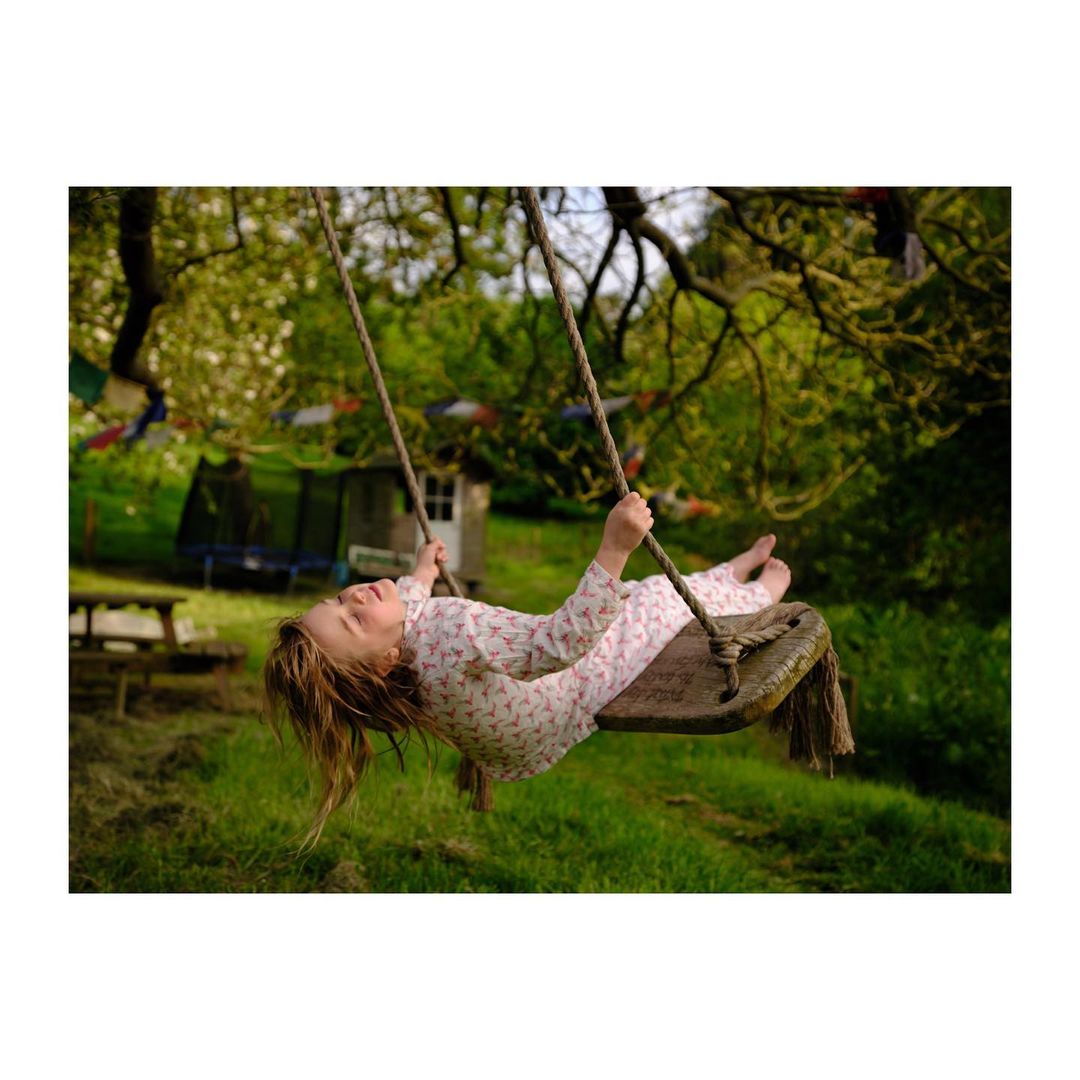A young girl swings on a wooden swing suspended from a tree in a grassy outdoor setting, with her hair hanging upside down.