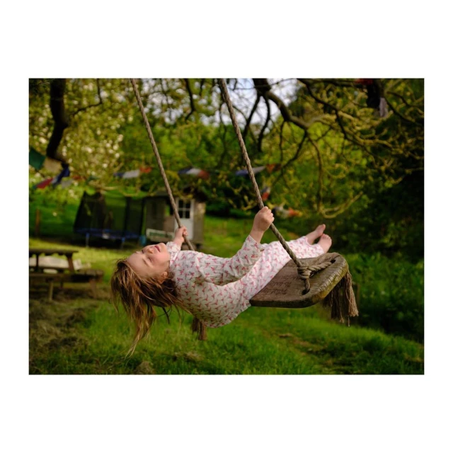 A young girl swings on a wooden swing suspended from a tree in a grassy outdoor setting, with her hair hanging upside down.