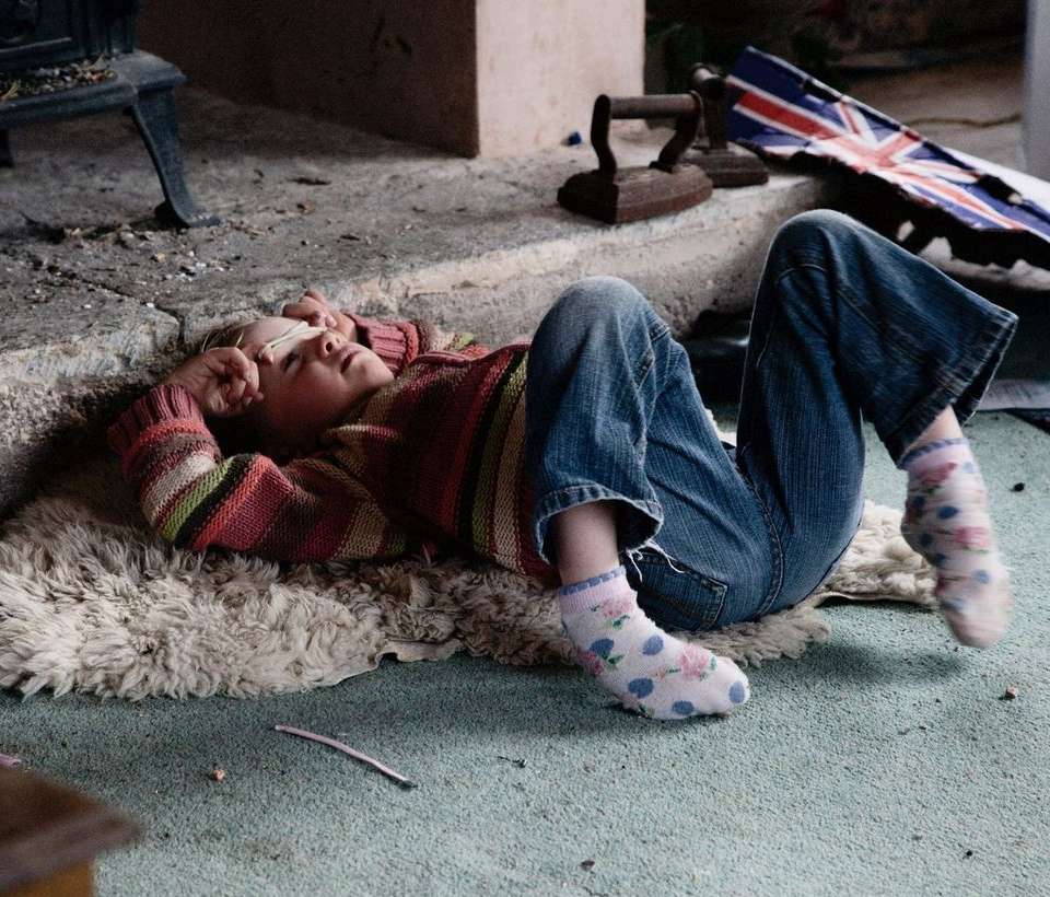 Young girl in a striped sweater and blue jeans lies on a sheepskin rug, in front of a dark fireplace, with a British flag visible.
