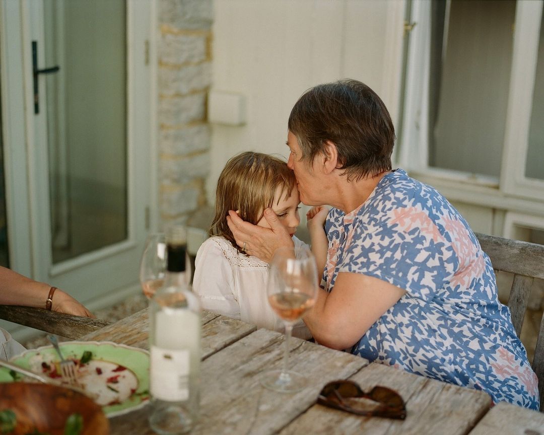 Grandmother lovingly kisses her granddaughter's forehead at an outdoor table setting with food and drinks, bathed in soft, natural light.