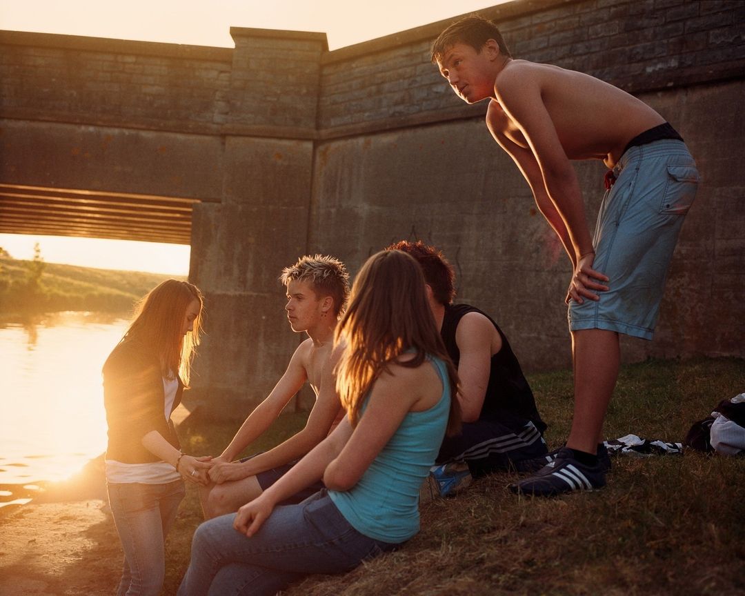 Group of teenagers gathered outdoors near a concrete structure and water, backlit by golden sunlight, creating a warm and casual atmosphere.