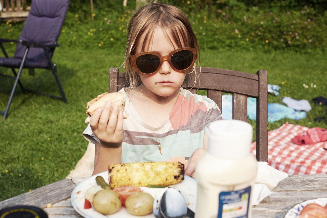 Child with sunglasses eating sandwich at wooden picnic table outdoors in sunny backyard. Lawn chair, grass, and blanket are blurred.