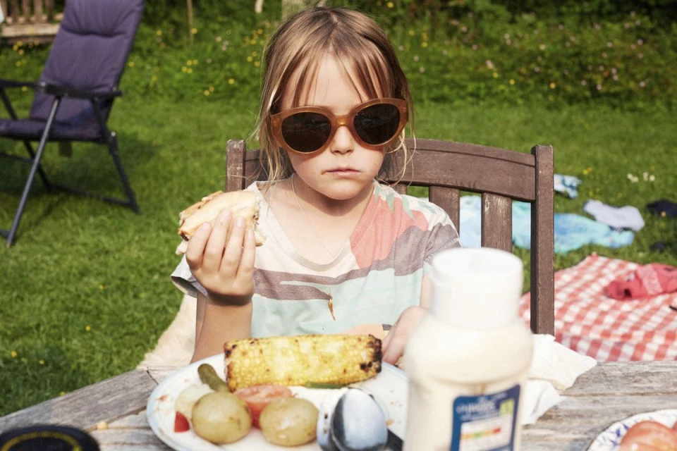 Child with sunglasses eating sandwich at wooden picnic table outdoors in sunny backyard. Lawn chair, grass, and blanket are blurred.