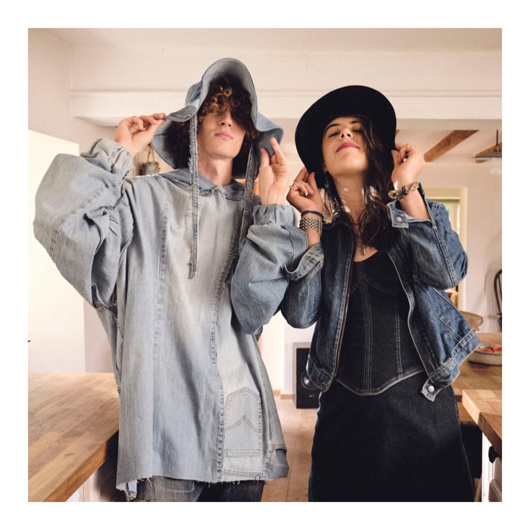Two people, a man and a woman, in an indoor setting, both wearing denim outfits. The woman wears a hat and the man a denim hooded shirt.