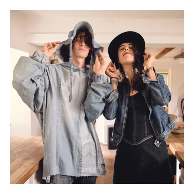 Two people, a man and a woman, in an indoor setting, both wearing denim outfits. The woman wears a hat and the man a denim hooded shirt.