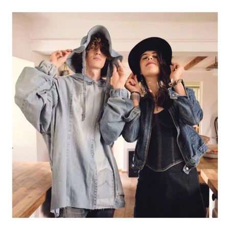 Two people, a man and a woman, in an indoor setting, both wearing denim outfits. The woman wears a hat and the man a denim hooded shirt.