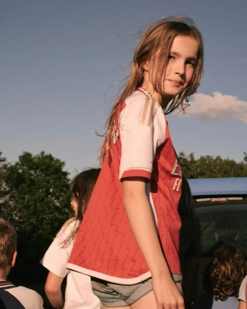 Girl in soccer jersey with group of children outside on sunny day. Girl in red and white soccer jersey looks at camera.