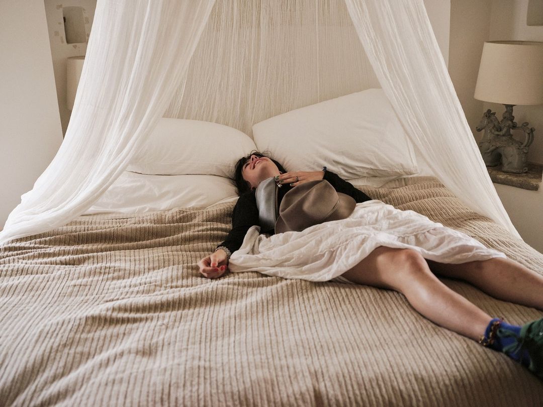 An adult woman is lying on a bed with white bedding and a beige cover, holding a hat in her hand in an indoor bedroom setting.