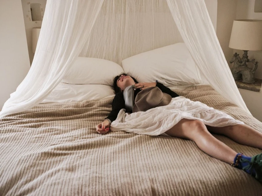 An adult woman is lying on a bed with white bedding and a beige cover, holding a hat in her hand in an indoor bedroom setting.