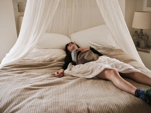 An adult woman is lying on a bed with white bedding and a beige cover, holding a hat in her hand in an indoor bedroom setting.