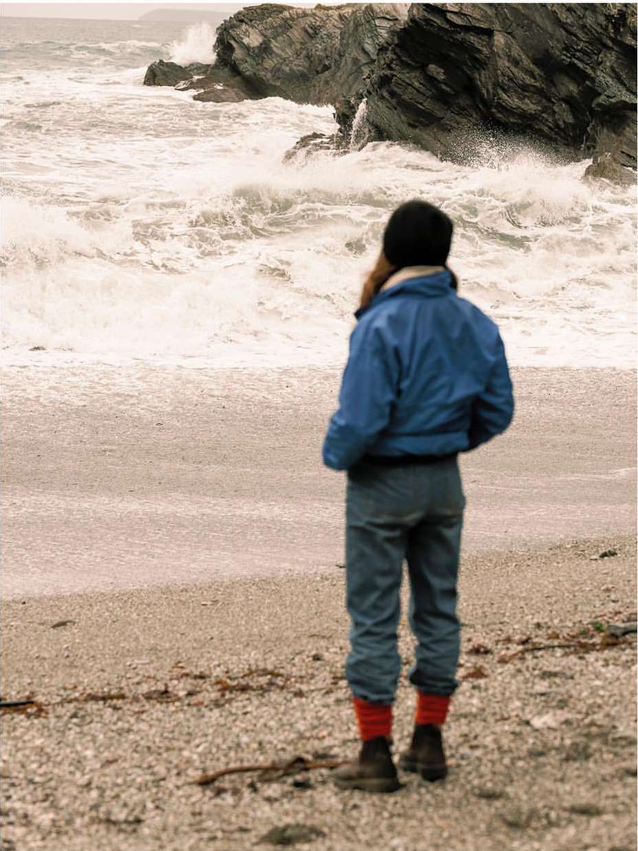 Person looks out at a stormy ocean with waves crashing against a rocky shore. The beach is sandy with rocks and driftwood.