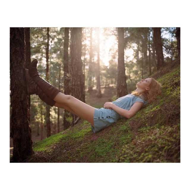 Woman in blue dress and brown boots relaxes against tree trunk in sunlit forest clearing with her legs elevated.