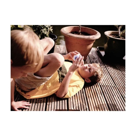 Two children playing outdoors on a wooden deck near potted plants. One child lies on their back while holding a toy.