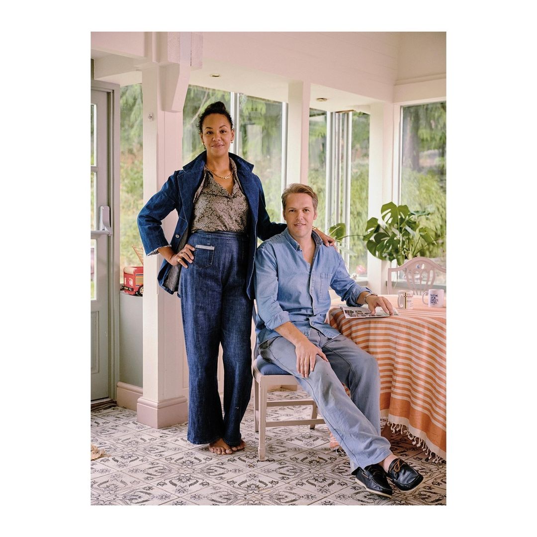 Portrait of a couple in a sunroom with casual clothing. Woman stands, man sits. Visible table, chair, houseplant, and large windows.