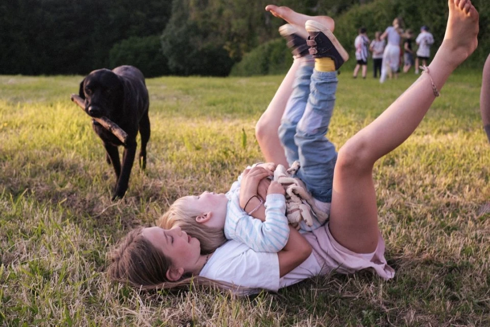Two children laying in a grassy field with a black dog in the background holding a stick, outdoor on a sunny day.