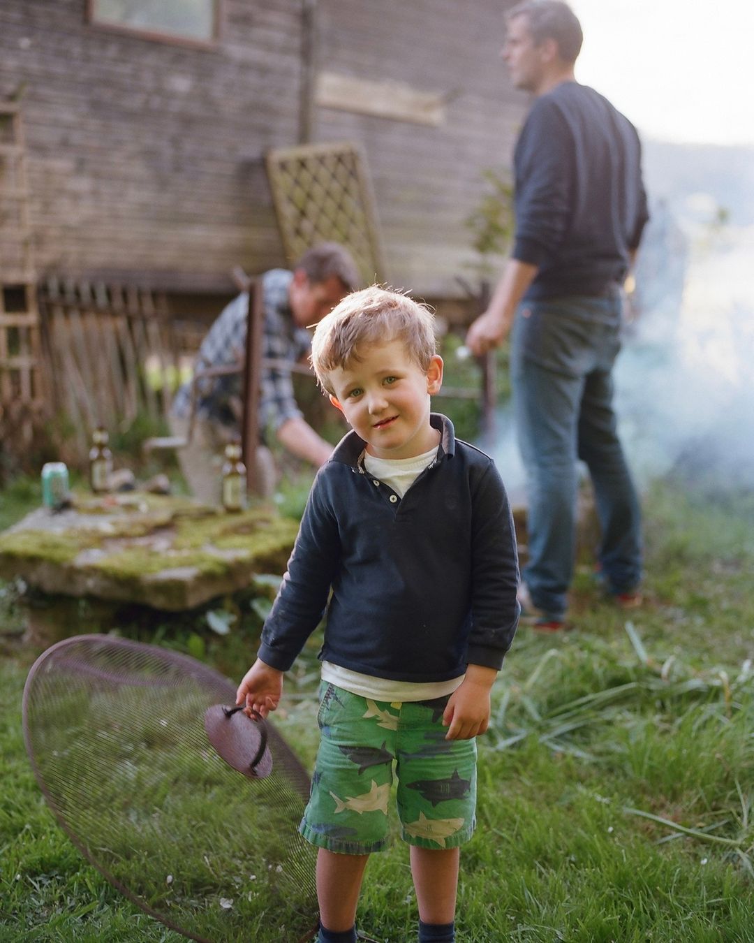 Young boy stands in backyard near grill, wearing green shark shorts, with two adult men visible in background during daytime.