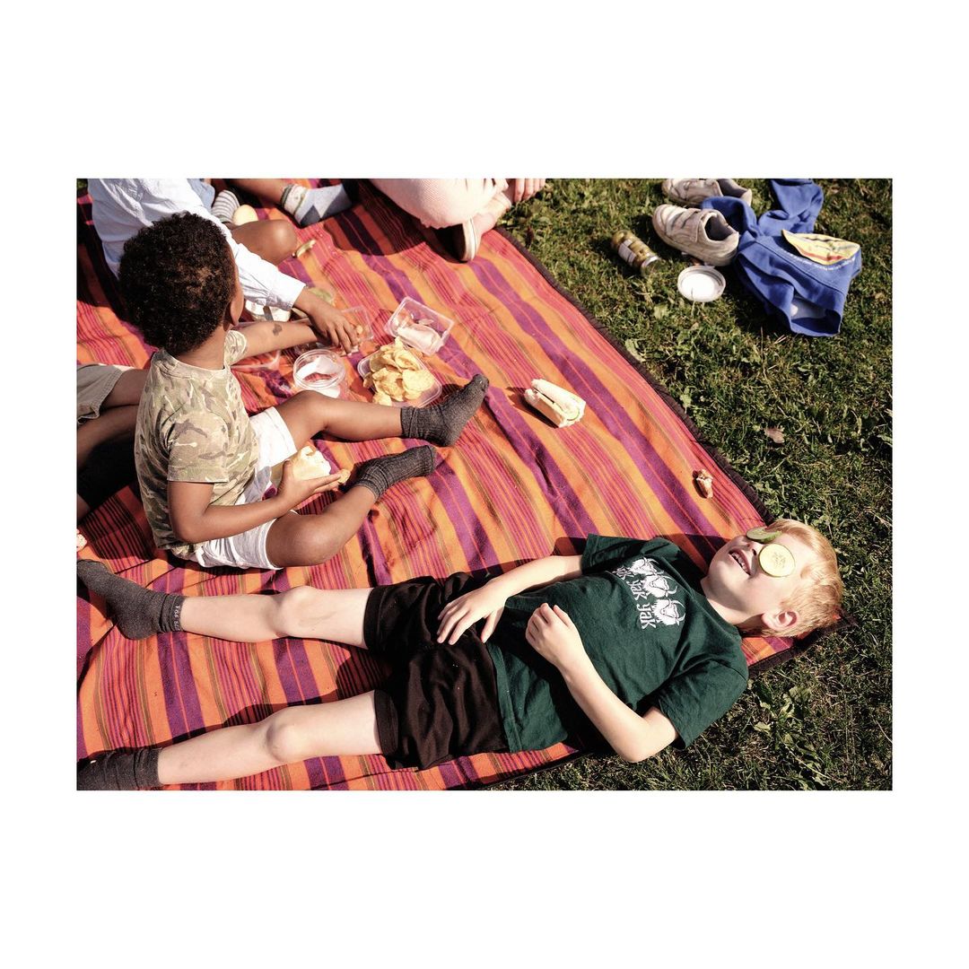 Children enjoy a sunny picnic on a striped blanket in a grassy outdoor setting, with one child relaxing with cucumber slices on eyes.